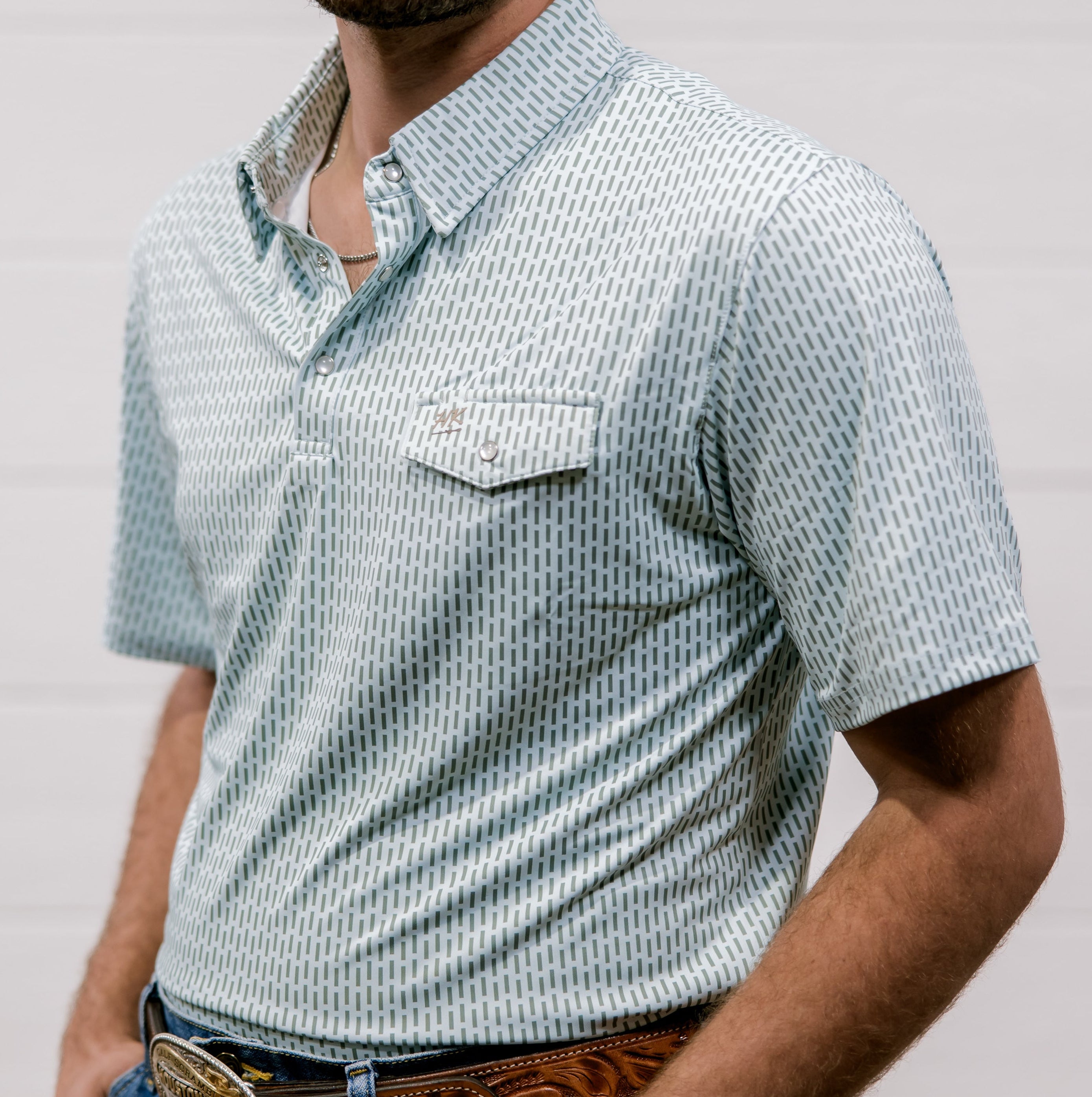 Man wearing a blue and green patterned shirt and jeans with a belt, standing against a plain background. side facing.