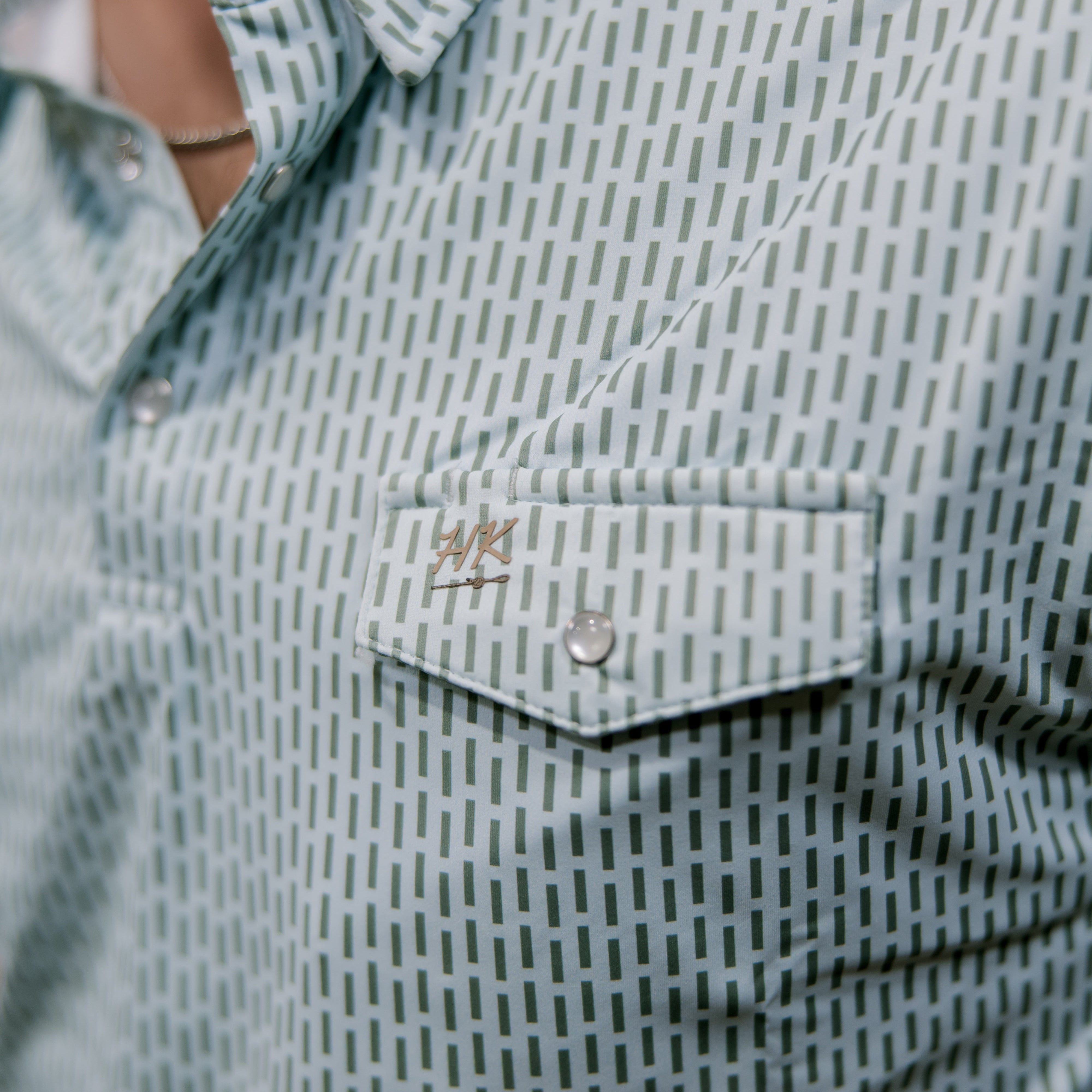 Close-up of a light blue and green patterned shirt with a brand logo on the chest pocket.