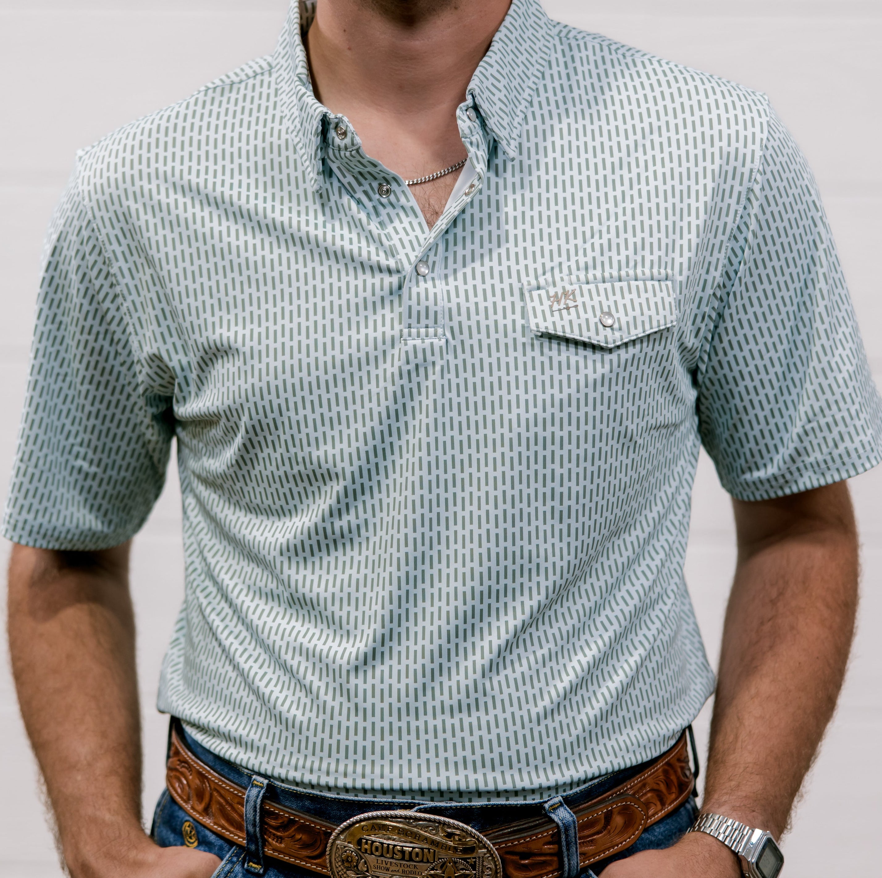 Man wearing a  blue and green patterned shirt and jeans with a belt against a plain background. front facing. 