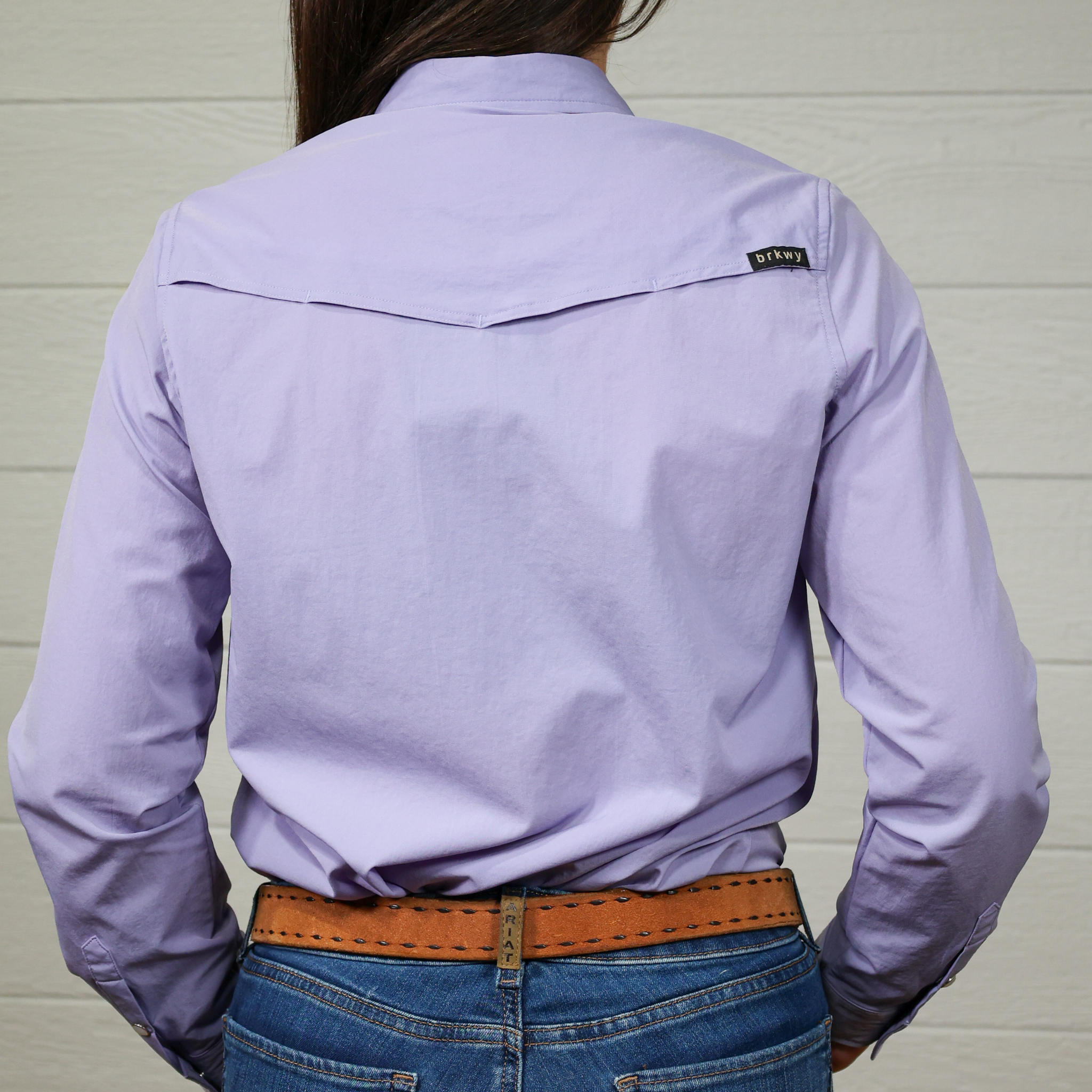 Person wearing a light purple shirt with a brand logo, standing against a plain white background